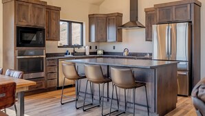 A modern kitchen with wooden cabinets, stainless steel appliances, a large island with bar stools, and a window providing natural light. - LaLuna Ranch- 4 bedroom mountain-view home on the way to Yellowstone (Cody)