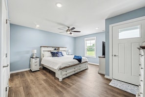 A bedroom with a bed, nightstands, a ceiling fan, a window, and a door. - Beach Glass Bungalow- new build close to beach and park (South Haven)