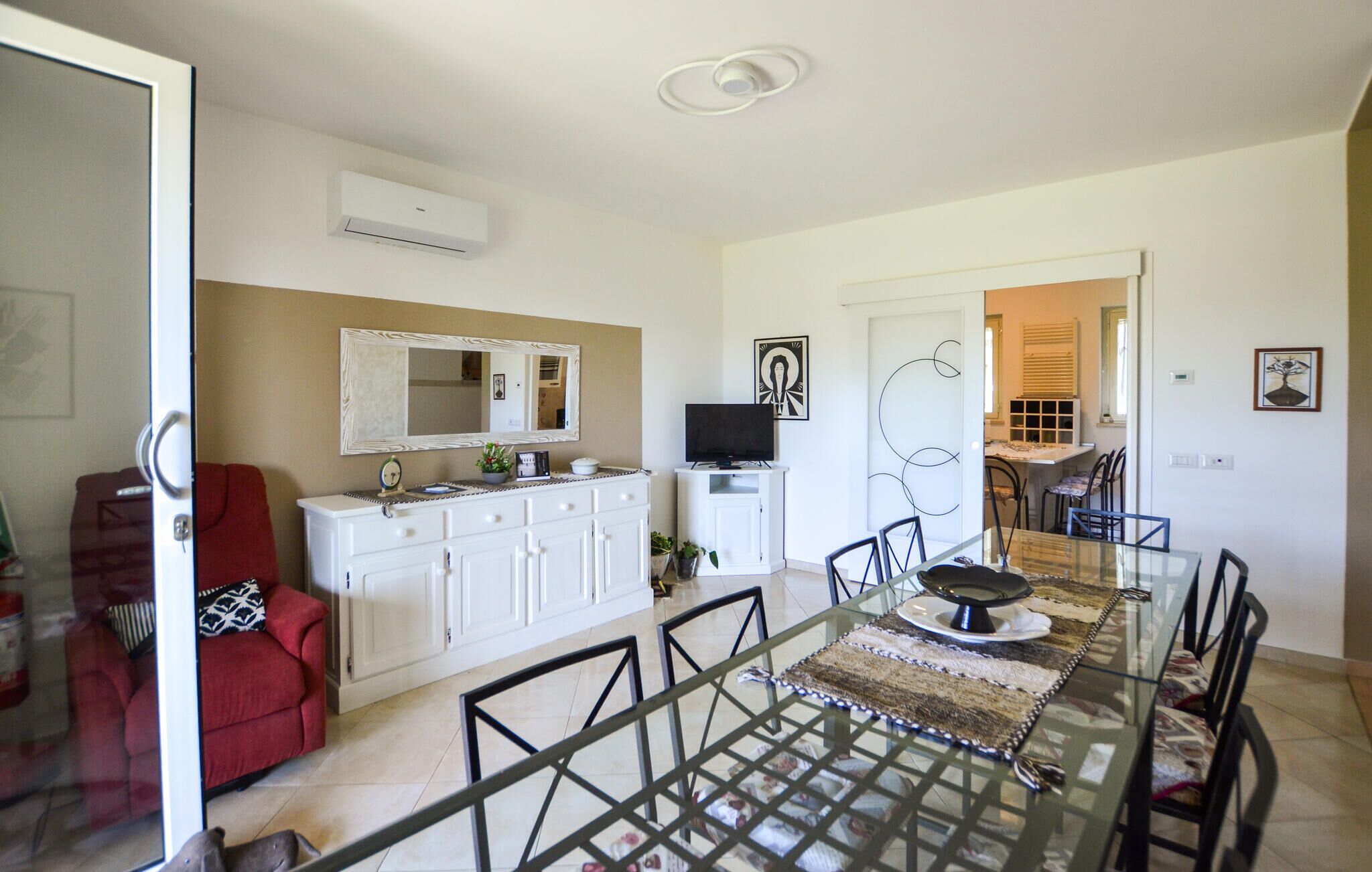 A dining area with a glass table, chairs, a white cabinet, a red chair, a TV, and a door leading to another room.