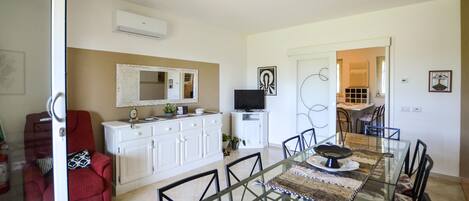 A dining area with a glass table, chairs, a white cabinet, a red chair, a TV, and a door leading to another room.
