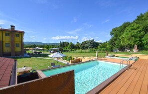 Ein Pool mit Holzterrasse, einem überdachten Sitzbereich und einem Gebäude mit gelber Fassade.