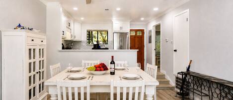 Un comedor con una mesa blanca dispuesta para cuatro personas, rodeada de sillas blancas, un ventilador de techo y vista a la cocina al fondo.