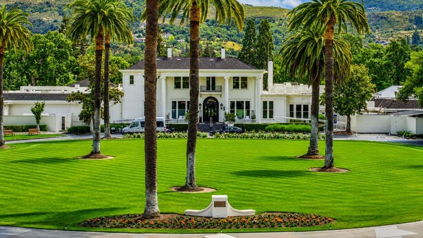 Una casa blanca de dos pisos con fachada simétrica, rodeada de un césped cuidado y palmeras, con un paisaje montañoso al fondo.