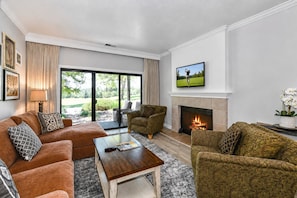 A living room with a fireplace, a television mounted above it, and a large window with a view of the outdoors.