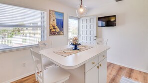 A dining area with a white table, chairs, a clear glass pendant light, a painting, and a TV on the wall. - Turtlemound Terrace - Your Favorite Beach Getaway (New Smyrna Beach)