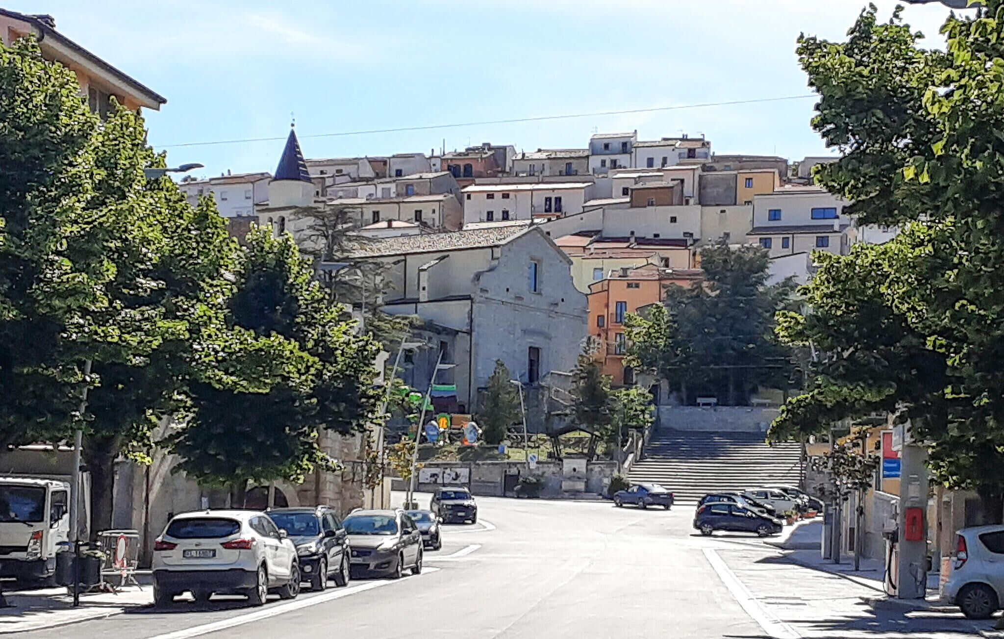 A street with parked cars, trees lining the road, and buildings in the background.