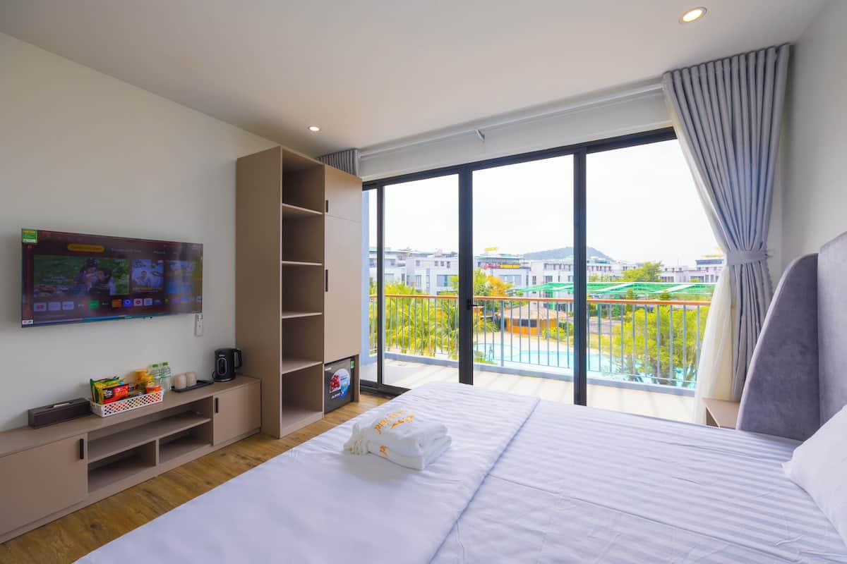 A modern hotel room with a large bed, a TV on the wall, a balcony with a view of buildings and greenery, and a shelf with items.