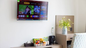 Una habitación de hotel con una televisión mostrando un servicio de streaming, una cafetera, un cesto de aperitivos, un florero con flores y una silla con estampado.