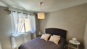 Ein Schlafzimmer mit einem großen Bett, einem Fenster mit durchsichtigen Vorhängen, einem Nachttisch mit Lampe und einem Heizkörper.