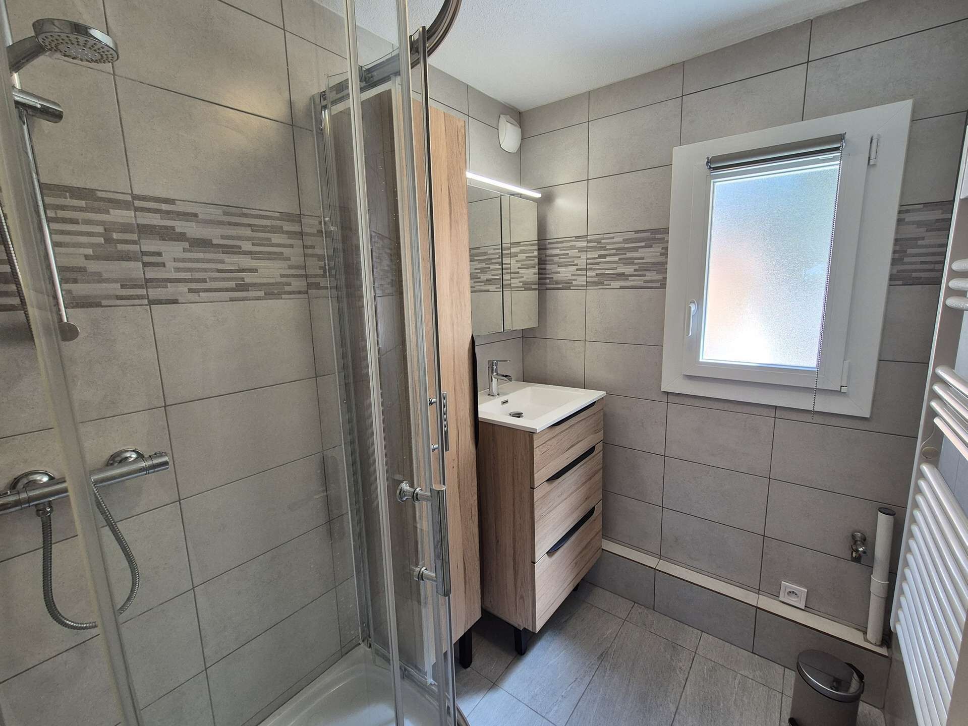 A modern bathroom with a glass shower enclosure, a wooden vanity, a mirror, and a window.