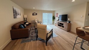 Una sala de estar con un sofá marrón, un sillón beige, un televisor sobre un mueble de madera y una zona de comedor con mesa y sillas.