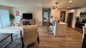 Una cocina moderna con una isla central, un comedor con sofá y mesa, y una sala de estar con televisor y plantas.