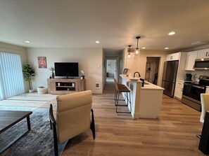 A modern kitchen with a central island, a dining area with a sofa and table, and a living room with a TV and plants.