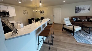 Una cocina moderna con encimera blanca, taburetes de bar, zona de comedor con sofá de cuero marrón y sala de estar con alfombra grande.
