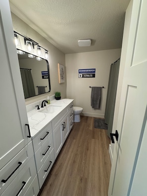 A bathroom with a white vanity, a sink, a toilet, a shower curtain, and a sign indicating the location.