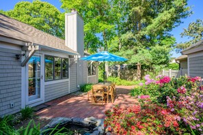 Ein Haus mit Terrasse, einem Holztisch und Stühlen, einem blauen Sonnenschirm und einem blühenden Garten.