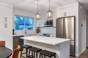 A modern kitchen with white cabinetry, stainless steel appliances, and a large island with bar stools. - Alpine Elegance | Designer Main Floor (Calgary)