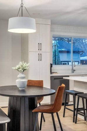 A modern kitchen with white cabinets, a round dining table with two chairs, and a hanging light fixture. - Alpine Elegance | Designer Main Floor (Calgary)