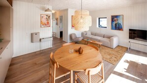 Una sala de estar moderna con una mesa redonda de madera, dos sillas de mimbre, un sofá color beige, un televisor y un ventanal grande.