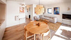 En moderne stue med et rundt træbord, to fletstole, en beige sofa, et fjernsyn og et stort vindue.