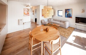 Ein modernes Wohnzimmer mit einem runden Holzesstisch, zwei Korbstühlen, einem beigen Sofa, einem Fernseher und einem großen Fenster.