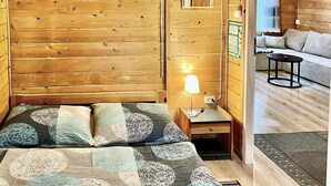 A wooden cabin interior with a bed, a nightstand, a lamp, and a sofa in the adjacent room.