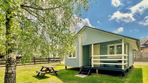 A small, light green wooden cabin with a porch and a picnic table in a grassy area.