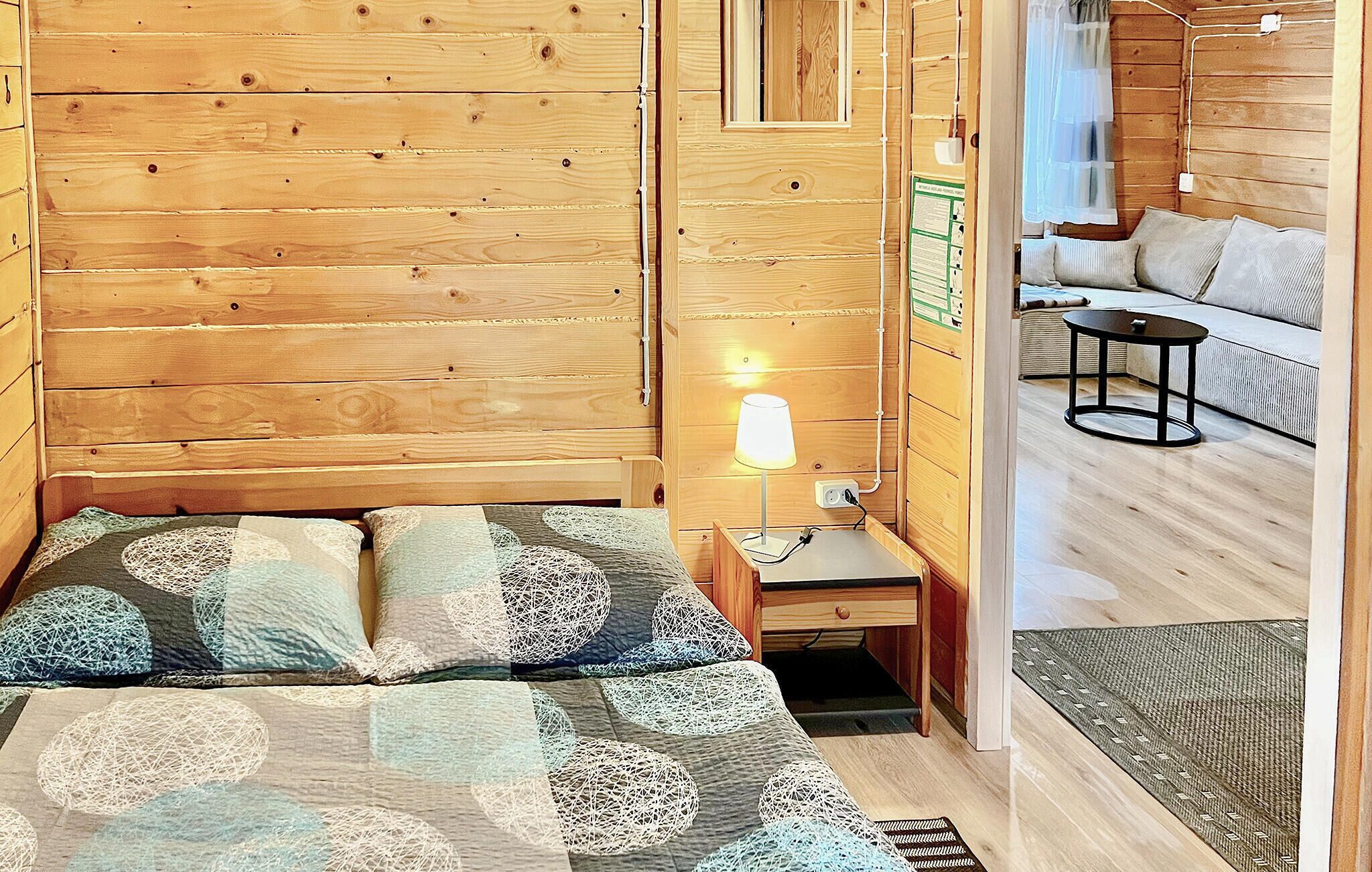 Une pièce en bois comprenant un lit, une table de chevet, une lampe et donnant sur une autre pièce où se trouvent un canapé et une table.