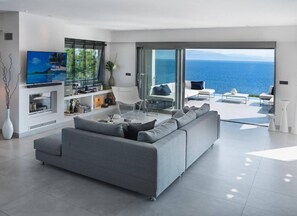 Ein modernes Wohnzimmer mit einem grauen Sofa, einem Flachbildfernseher und einem großen Fenster mit Meerblick.