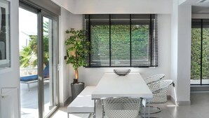 Ein moderner Essbereich mit einem weißen Tisch, Metallstühlen, einer Zimmerpflanze und einem Fenster mit Jalousien.