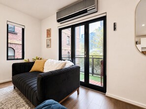 Ein modernes Wohnzimmer mit einem schwarzen Sofa, einem weißen Sitzkissen und einem großen Fenster mit Blick auf ein Backsteingebäude und Grünflächen.