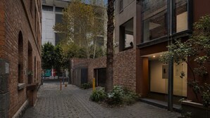 Un stretto vicolo acciottolato con edifici in mattoni, un piccolo albero e l'ingresso di un palazzo con una porta in vetro.