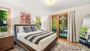 Un dormitorio con cama, mesitas de noche, un cuadro, una ventana y una puerta corredera de vidrio que da a un balcón.