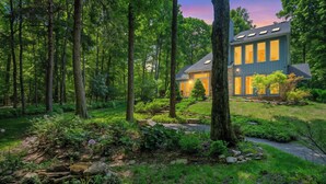 A modern house surrounded by a forested area with a well-maintained garden and a stone pathway leading to the entrance.