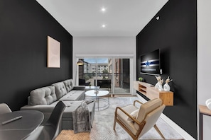 A modern living room with a grey sofa, a coffee table, a TV mounted on the wall, and a balcony with a view of buildings. - ToyotaStadium+Gym+Pool+Golf+W+D (Frisco)