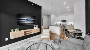 Ett modernt vardagsrum med en platt-TV, en grå soffa, ett soffbord i trä och ett kök med vita skåp och en köksö.