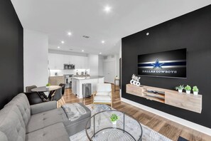 A modern living room with a grey sofa, a TV mounted on a black wall, and a kitchen in the background. - Gym+Pool+FriscoSquare+ToyotaStadium+PGAGolf+W+D (Frisco)