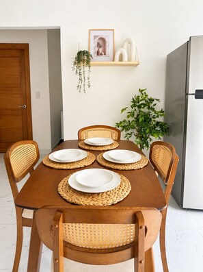 Ein Esstisch mit vier Tellern, Korbstühle, ein an der Wand befestigtes Regal mit Dekoration und ein Kühlschrank.