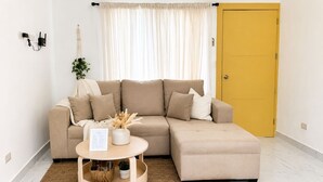 En stue med en beige modulsofa, et lille rundt sofabord med en plante, en gul dør og en hvid væg med et stikkontakt.