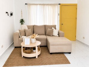 Ein Wohnzimmer mit einem beigefarbenen Ecksofa, einem kleinen runden Couchtisch mit einer Pflanze, einer gelben Tür und einer weißen Wand mit einer Steckdose.
