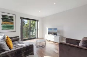 Ein modernes Wohnzimmer mit einer Couch, einem Couchtisch, einem Fernseher auf einem weißen Schrank und einem großen Fenster mit Blick auf Grünflächen.