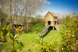 Ein hölzernes Spielhaus mit Rutsche in einer grünen Rasenfläche mit Bäumen.