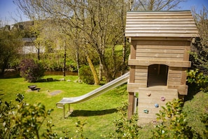 Ein hölzernes Spielhaus mit Rutsche und Kletterwand, umgeben von Grünpflanzen und Bäumen.