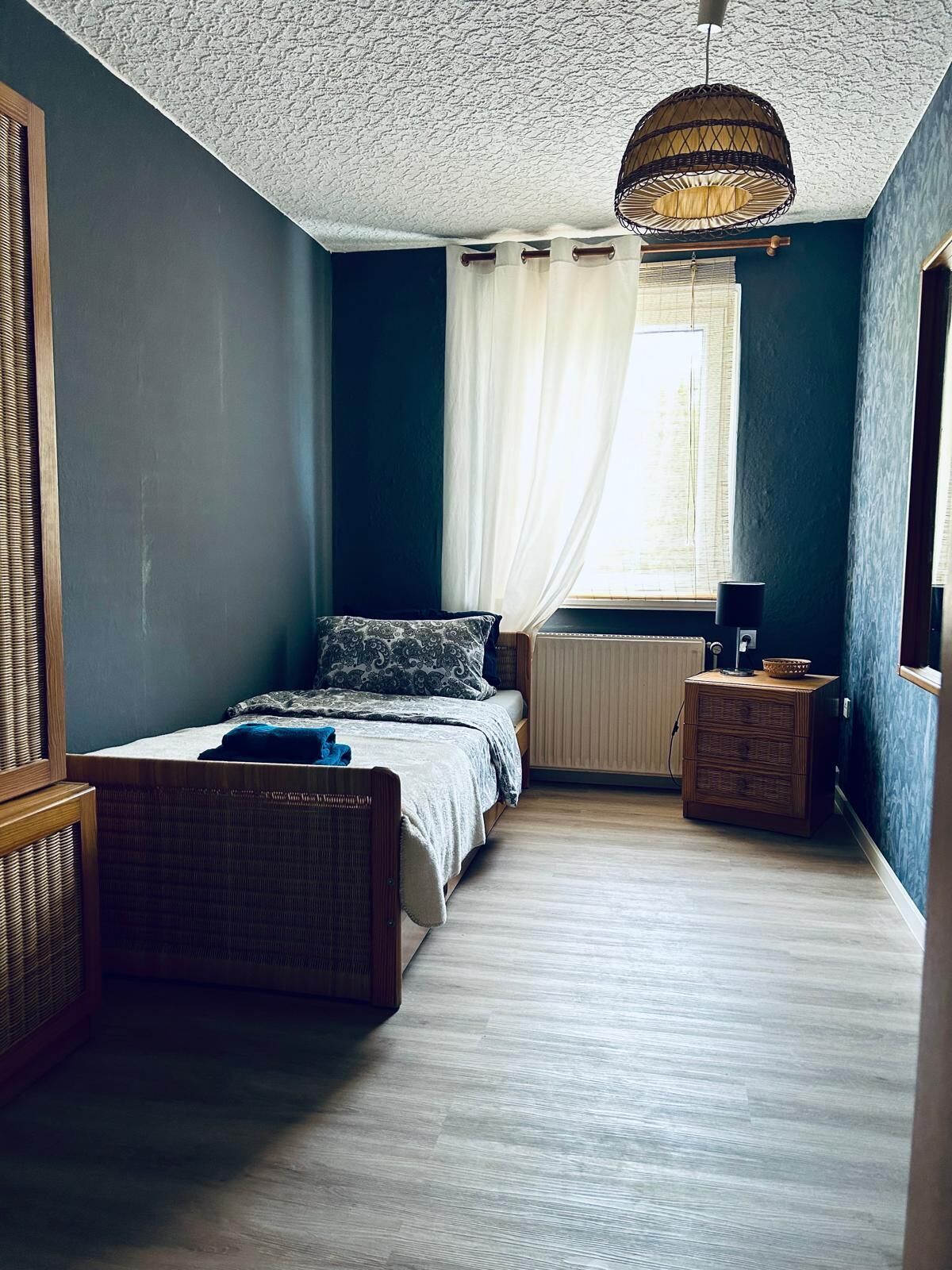 A bedroom with a bed, a bedside table, a radiator, a window with curtains, and a hanging light fixture.