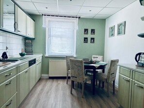 Une cuisine équipée d’une table à manger, de chaises, de placards, d’une fenêtre avec des stores et de tableaux accrochés au mur.