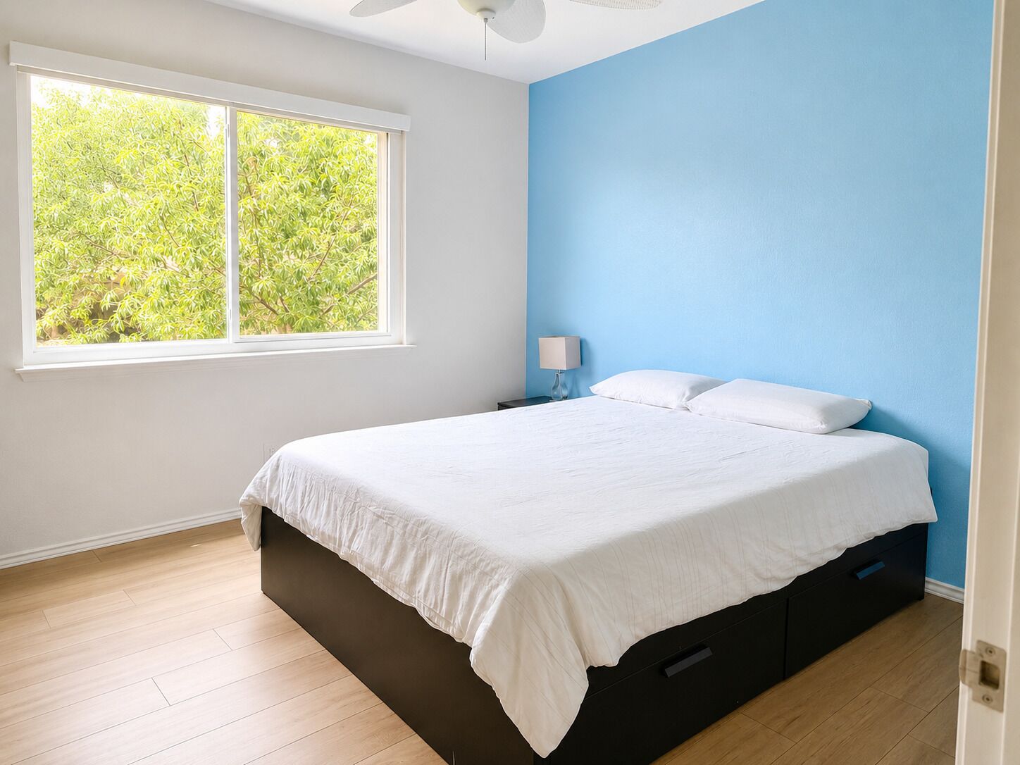 Une chambre avec une grande fenêtre, un lit, une table de chevet et un mur d’accent bleu.