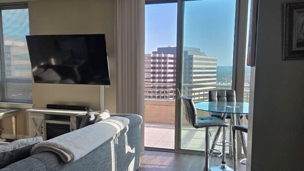 Een moderne hotelkamer met een bank, een flatscreen-tv, een zithoek met een glazen tafel en stoelen, en een grote schuifpui naar een buitenbalkon.