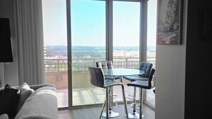 Een moderne hotelkamer met een groot schuifglas dat uitkomt op een balkon, een glazen eettafel met stoelen, een bank en een schilderij aan de muur.
