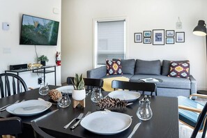 Ein modernes Wohnzimmer mit einem grauen Sofa, einem Esstisch für zwei Personen und einem Wand-Fernseher.