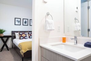 Ein modernes Hotelzimmer mit einem Bett, einem Nachttisch mit Pflanze, einem großen Spiegel über dem Waschbecken und einem an der Wand befestigten Handtuchhalter.